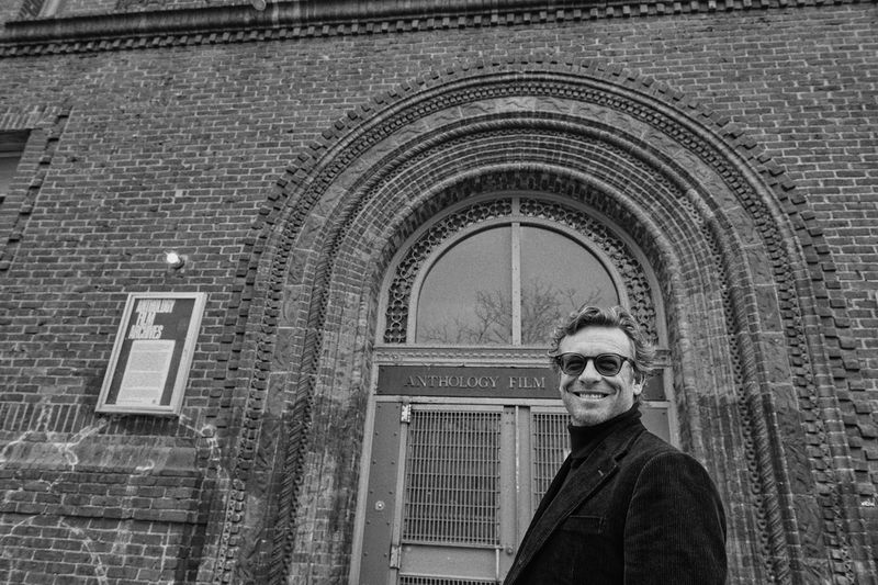 Black and white portrait of Simon Baker smiling outdoors in front of Anthology Film Archives building with an arched doorway and brick facade.