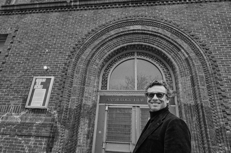 Black and white portrait of Simon Baker smiling outdoors in front of Anthology Film Archives building with an arched doorway and brick facade.