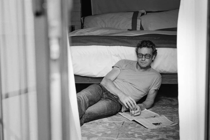Monochrome portrait of a man with glasses reclining indoors, reading a newspaper, with a bed and curtains visible in the background.