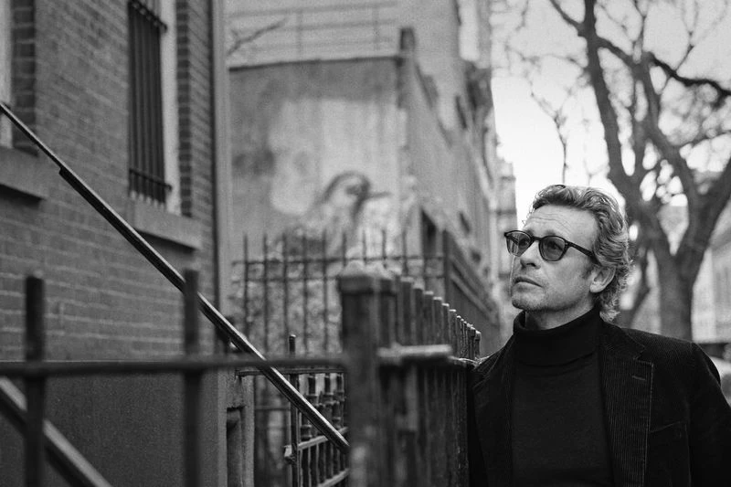 Black and white portrait of an adult male with glasses and dark turtleneck outdoors in front of a brick building and wrought iron fence.