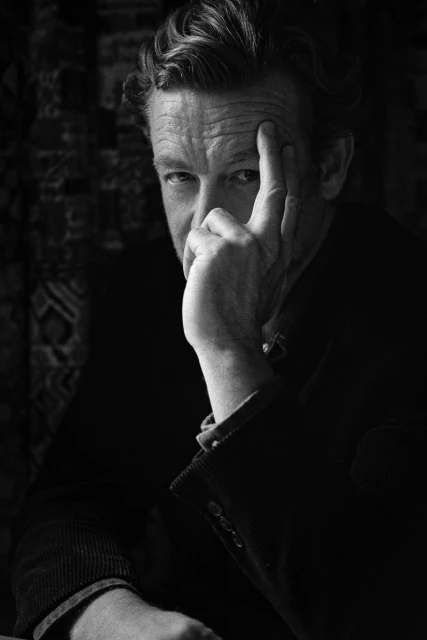 Black and white portrait of a mature man with curly hair in a dark coat, gazing intensely with his hand near his face.