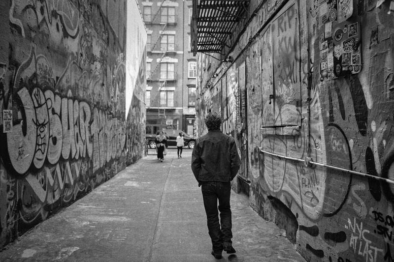 Black-and-white shot of a man walking down an alleyway with graffiti-covered walls, with two distant figures and a building in the background.