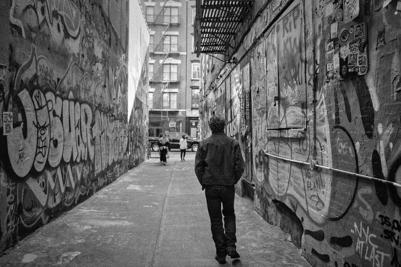 Black-and-white shot of a man walking down an alleyway with graffiti-covered walls, with two distant figures and a building in the background.
