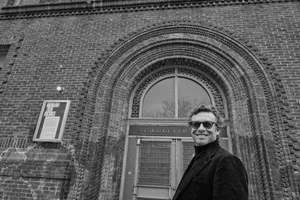 Black and white portrait of actor Simon Baker wearing sunglasses and dark jacket standing in front of brick building