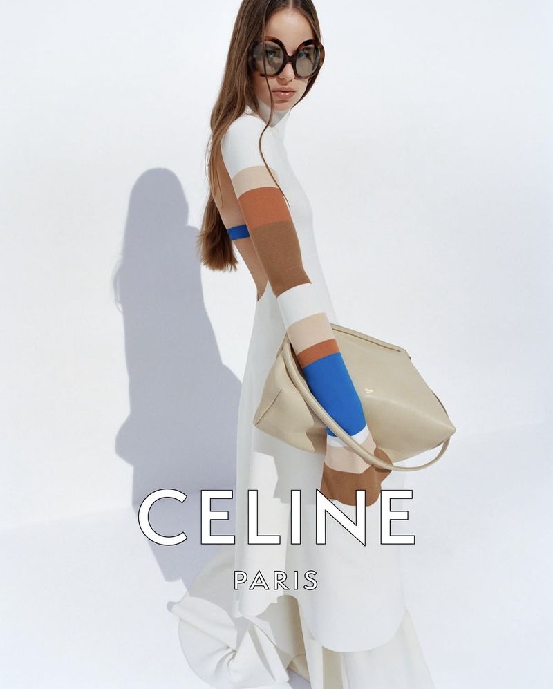 A woman with long brown hair in sunglasses poses in a white dress and beige purse on a white background for a fashion advertisement.