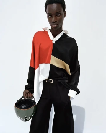 A fashion portrait features a Black woman with short hair wearing a color-block top and black trousers, holding a CELINE helmet against a white background.