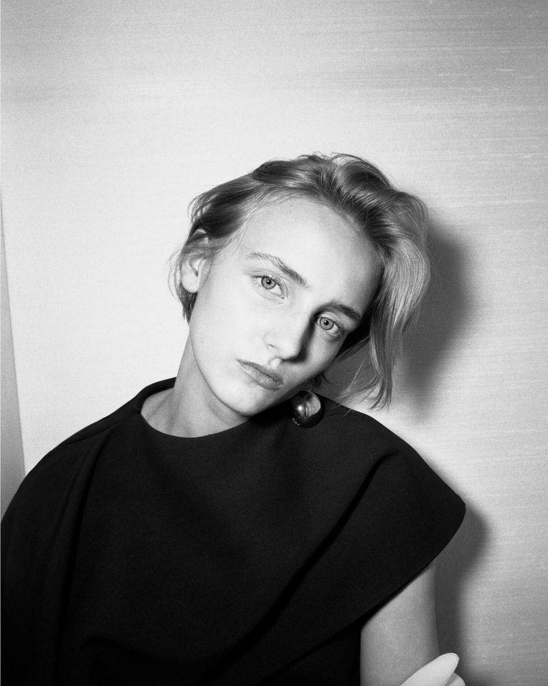 Black and white portrait of a light-skinned young woman with short brown hair, wearing a black top and hoop earring, against a plain backdrop.