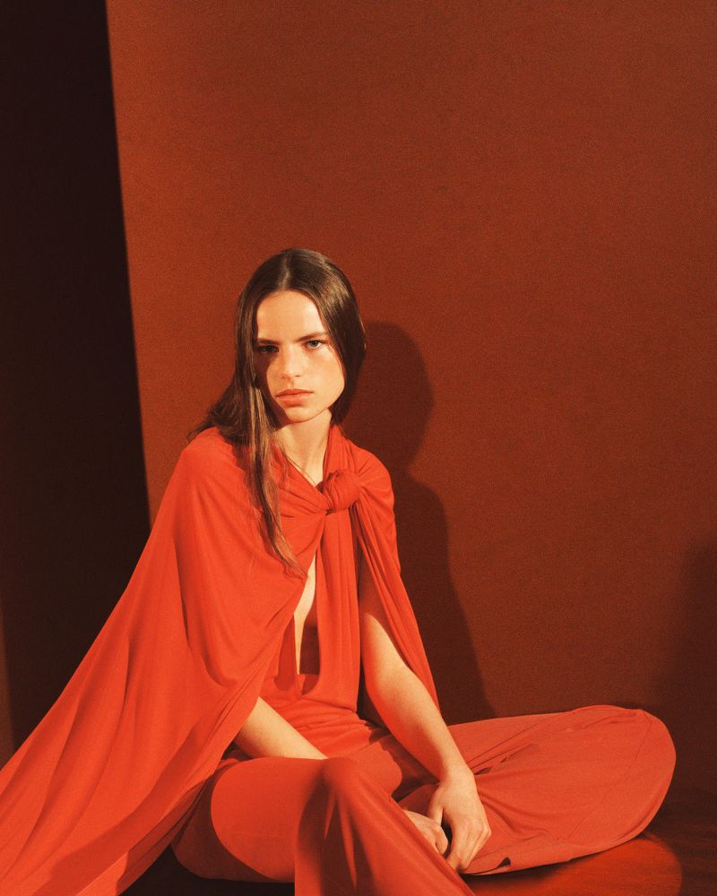 A woman in a red dress and cape sits indoors against a warm-toned wall. Studio lighting highlights the flowing fabric and her neutral expression.