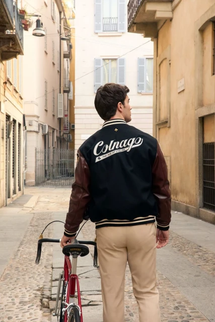 Man in jacket with bicycle stands in an outdoor alleyway, buildings on both sides; light colored pavement, back view.
