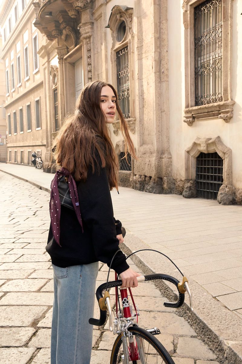 Woman with long brown hair, wearing a black sweatshirt and jeans, walking a vintage bicycle on a stone street with buildings.