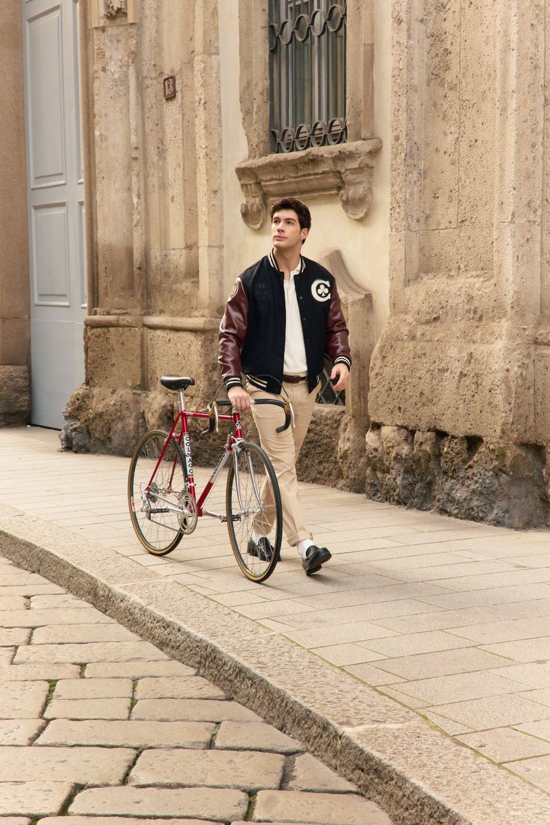A young man walks next to his red bicycle on a brick street in front of a stone building with decorative architectural details.