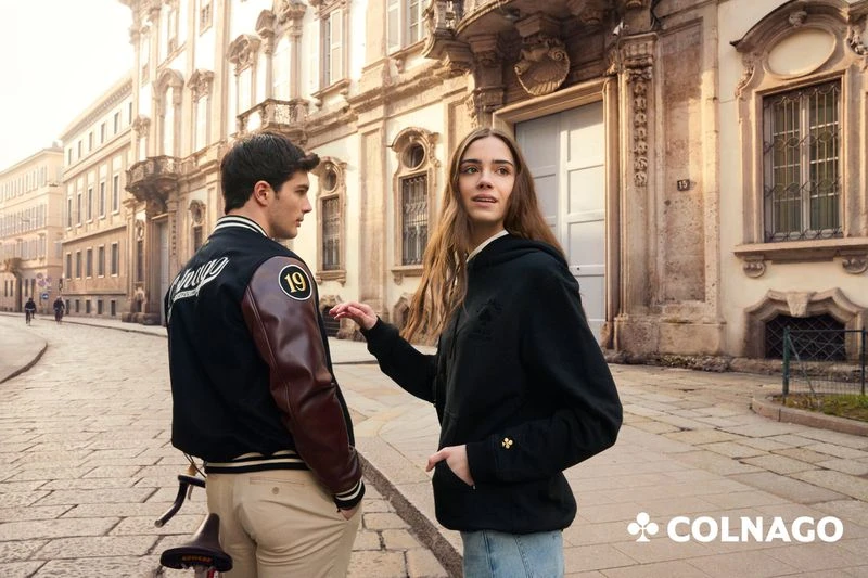 A young couple poses in an urban setting, with a man in a letterman jacket and a woman in a hoodie, against a backdrop of classic architecture.