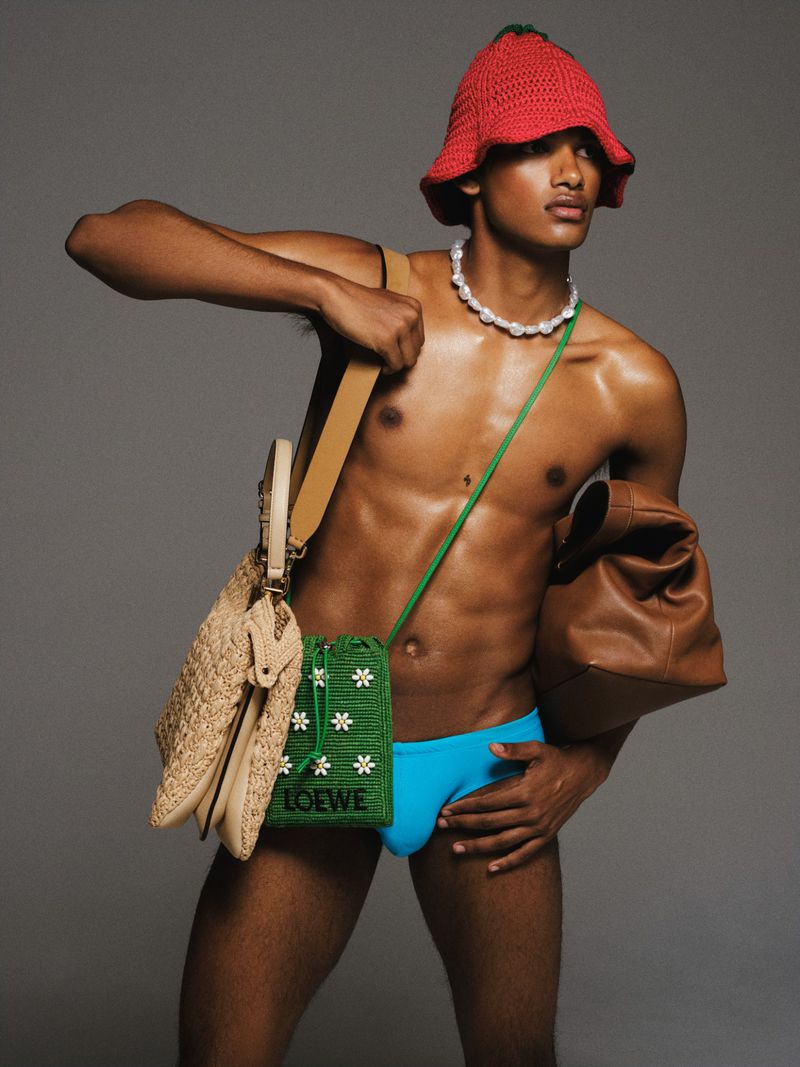 Dark-skinned male model wears a red crochet hat, swimsuit, and pearl necklace while holding purses in a grey studio setting.