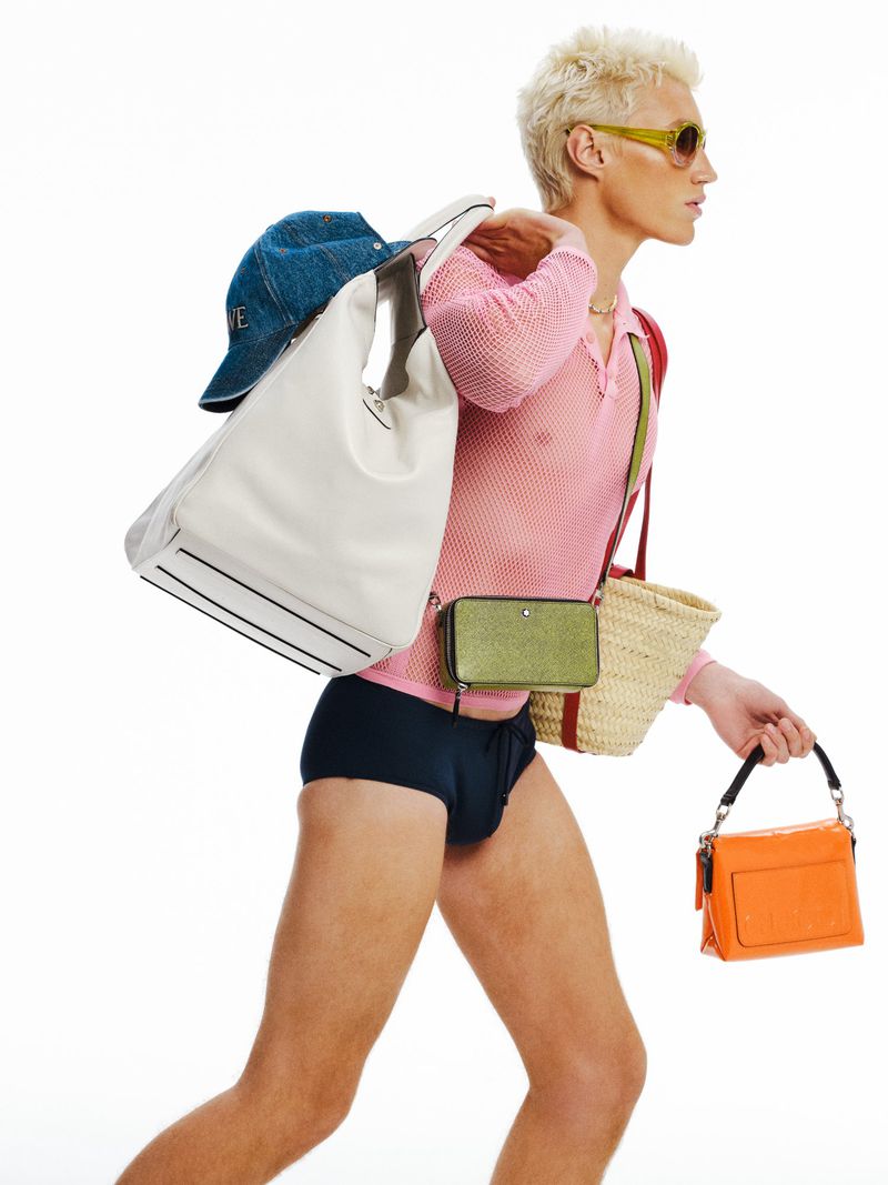 Fashionable person with blond hair carries multiple bags in a studio shot, wearing a pink shirt, dark shorts, and sunglasses.