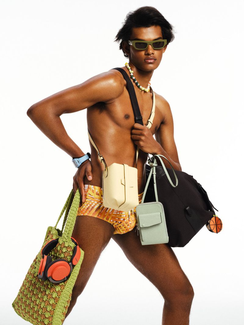 Male fashion model posing with an assortment of bags in a bright, studio setting wearing sunglasses and beaded necklaces.