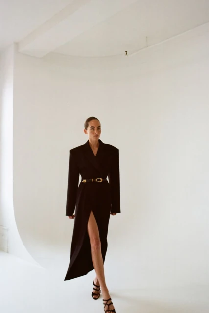 Woman in black blazer dress with gold belt poses in a bright white studio setting, showing a confident and stylish look.