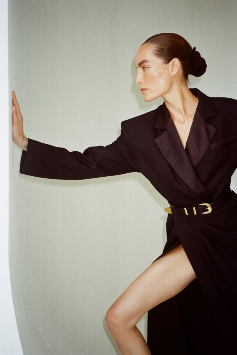 Fashion shot of a model wearing a black blazer dress with a gold belt, standing in front of a light grey wall in a photo studio.