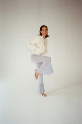 Model wearing a white jacket and sheer pleated trousers posing against a plain white studio background