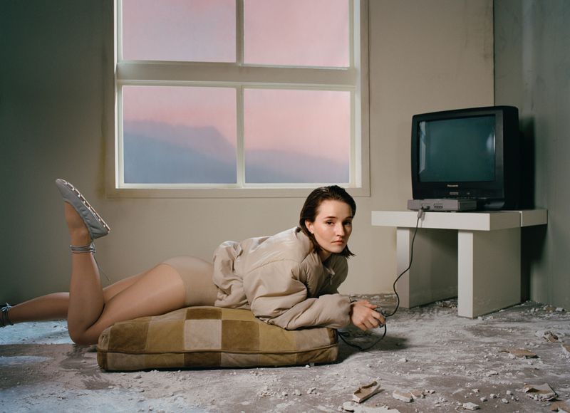 A young woman in casual clothes and a light jacket lies on a cushion with a vintage television in an indoor, minimalist setting.