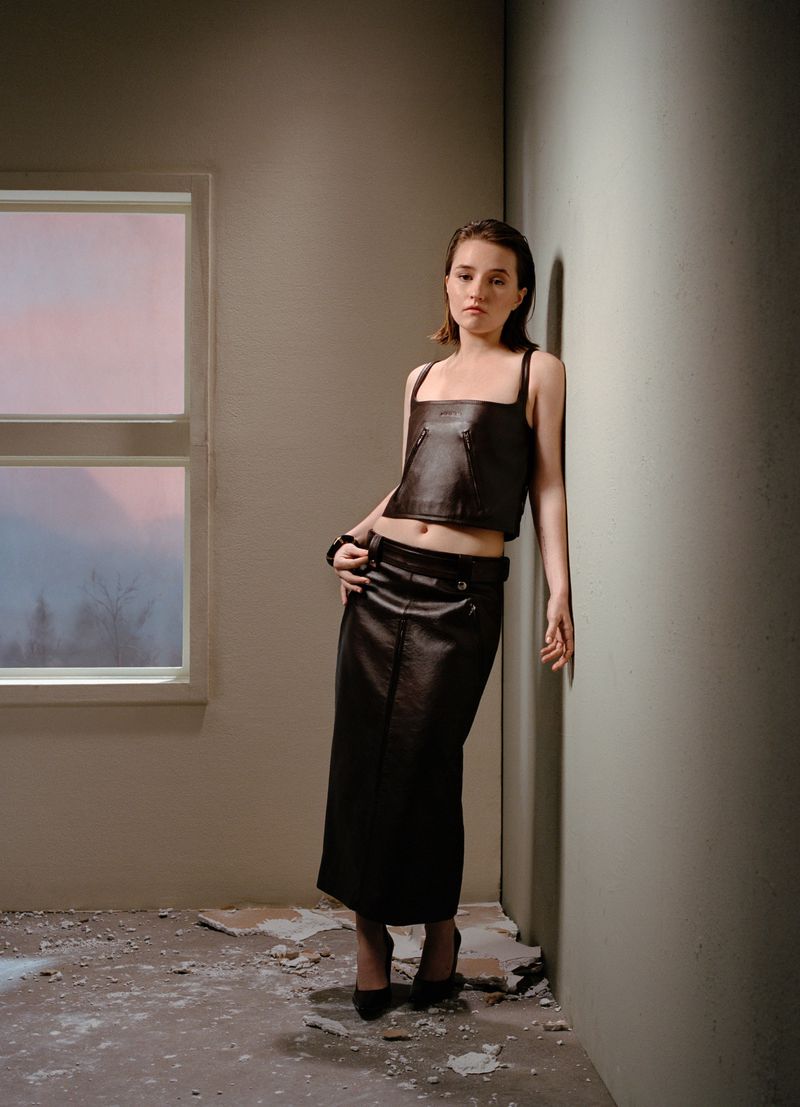 A young adult woman leans against a wall in an empty room. She is wearing a dark brown, two-piece leather outfit and black high heels.