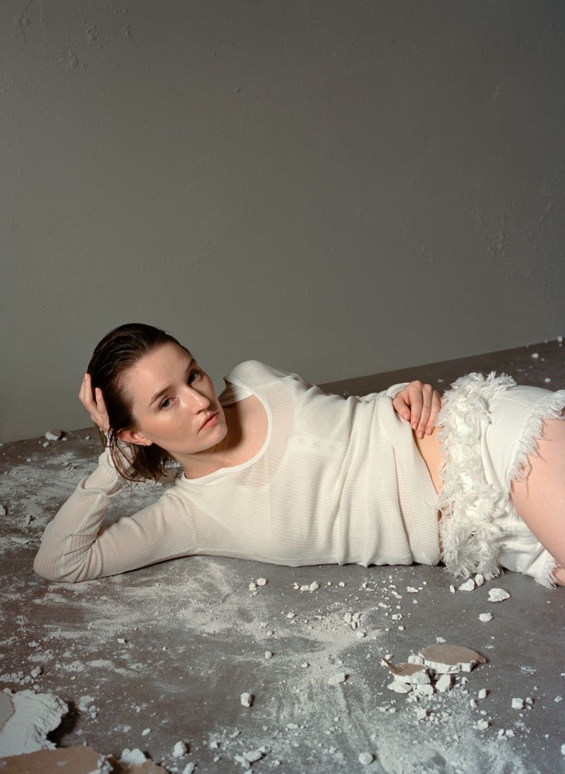 A young woman with wet hair poses while laying on her side wearing a white top and shorts in a grey room with debris on the floor.