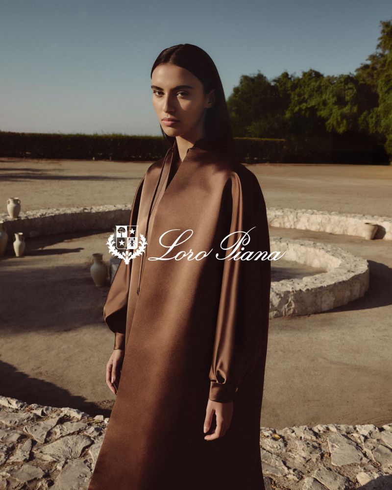 Woman in brown dress poses in outdoor fashion shot with villa and greenery in the background; Loro Piana logo visible.