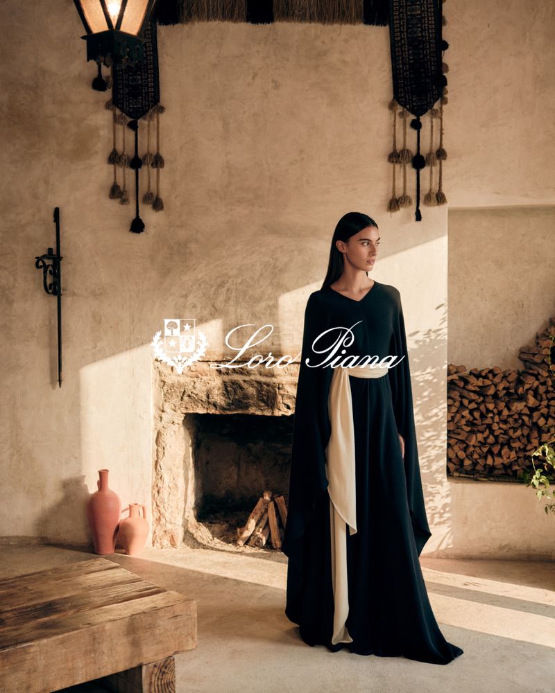 Woman in a long black dress with a cream sash poses in front of a stone fireplace in a room with neutral, rustic decor.
