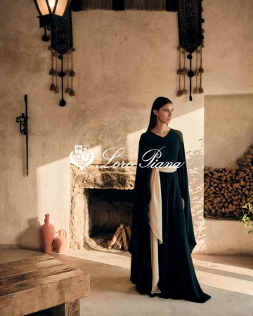 Woman in a long black dress with a cream sash poses in front of a stone fireplace in a room with neutral, rustic decor.