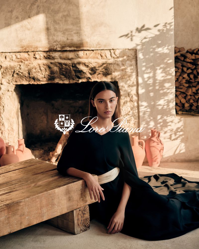Elegant woman poses in black dress with a white belt next to a rough-hewn table and a stone fireplace in an indoor setting with warm lighting.