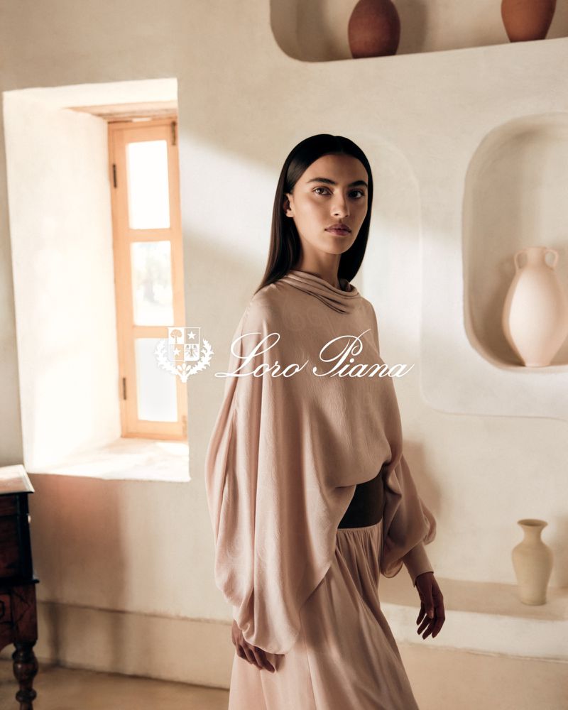 Elegant woman in a blush-toned outfit stands in a sunlit room with neutral walls and arched niches in a fashion advertisement for Loro Piana.
