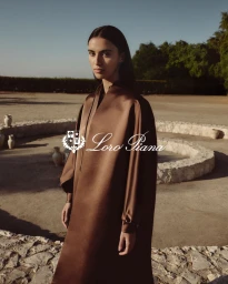 Model in a brown silk coat standing outdoors near a stone courtyard under a clear sky for Loro Piana Ramadan 2026 campaign.