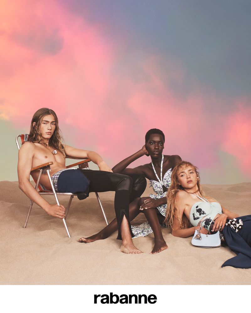 Three fashion models pose on a sandy surface against a colorful sky backdrop, wearing swimsuits and accessories in a fashion editorial shoot.