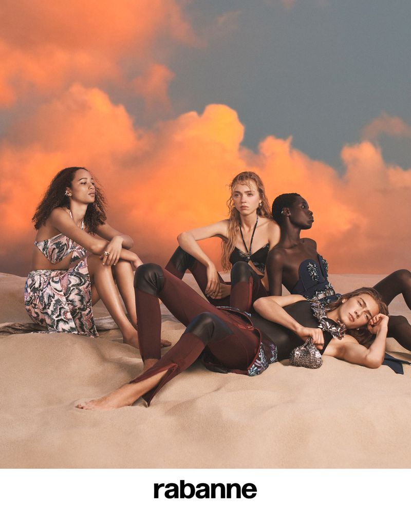 Group of fashion models posing on a sandy surface, wearing fashionable clothing with the rabanne logo visible, and a dramatic sky background.
