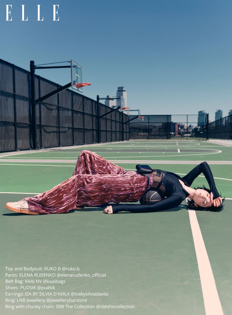 Fashion editorial shot of a model lying on a tennis court wearing a black top, velvet pants, and shoes, with a basketball next to her.