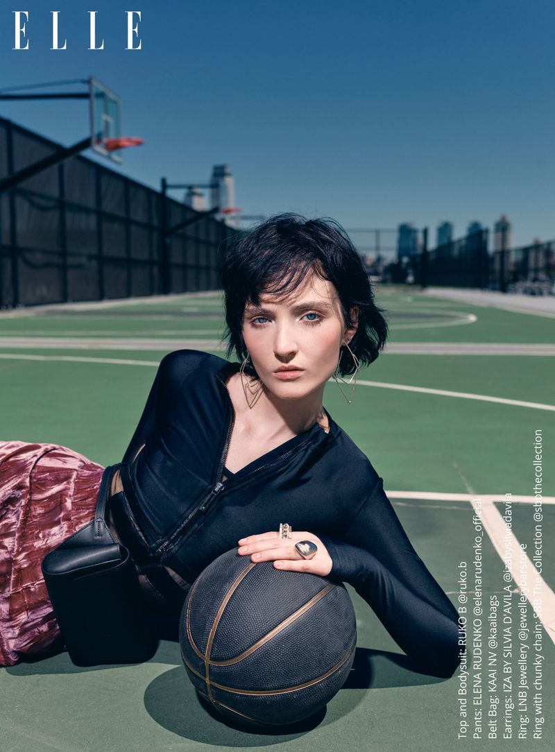 A young woman lies on a basketball court with a basketball, wearing a dark top, patterned pants, with buildings and blue sky background.