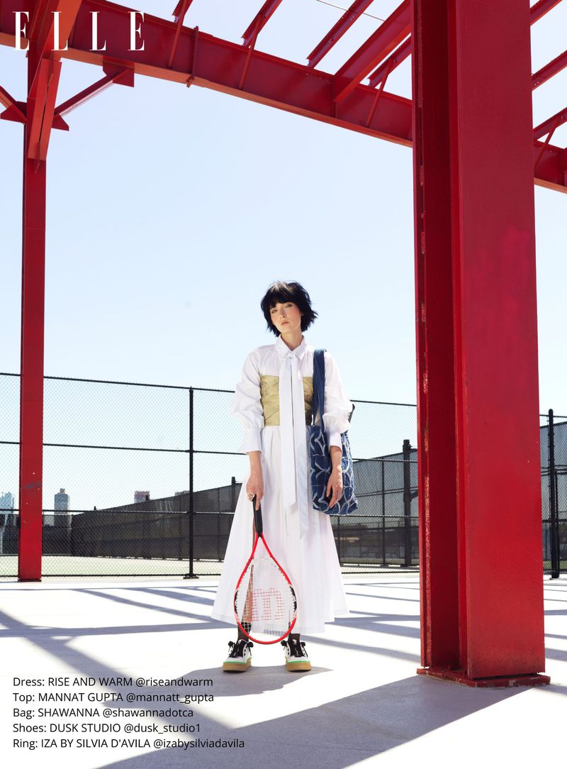 Young woman in a white dress and tennis shoes stands on a tennis court holding a racket under a red metal structure.