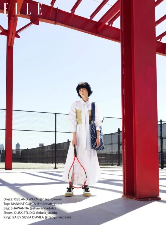 Young woman in a white dress and tennis shoes stands on a tennis court holding a racket under a red metal structure.