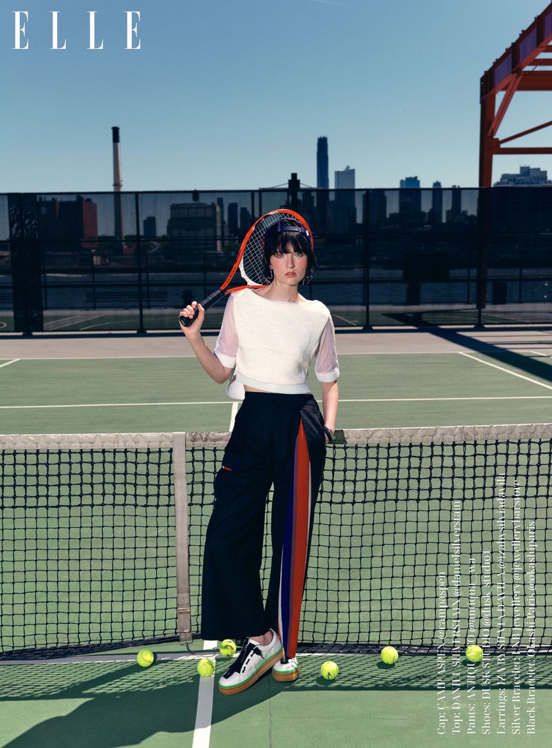 Fashion shot of a young adult woman in sportswear holding a tennis racket on a court, with a cityscape and 'ELLE' logo visible.