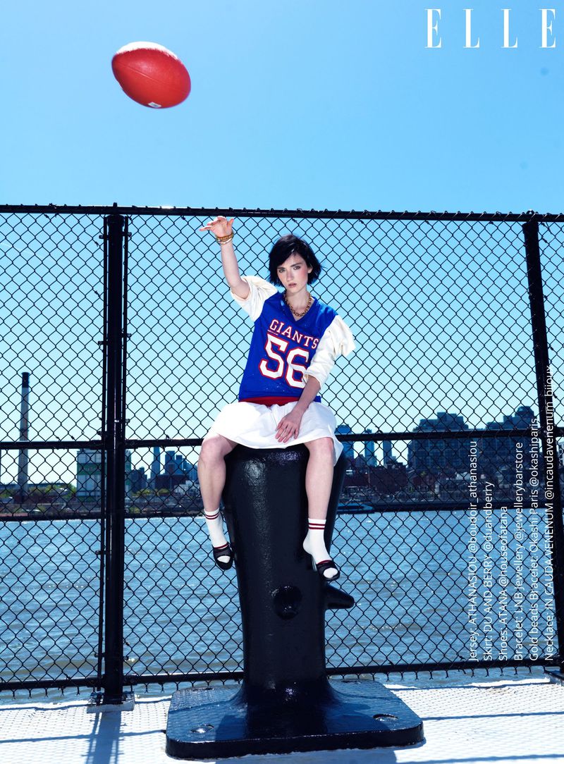 A young woman poses in a sports jersey with a football outdoors on a city pier in bright daylight with an azure sky. Elle magazine visible.