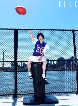A young woman poses in a sports jersey with a football outdoors on a city pier in bright daylight with an azure sky. Elle magazine visible.