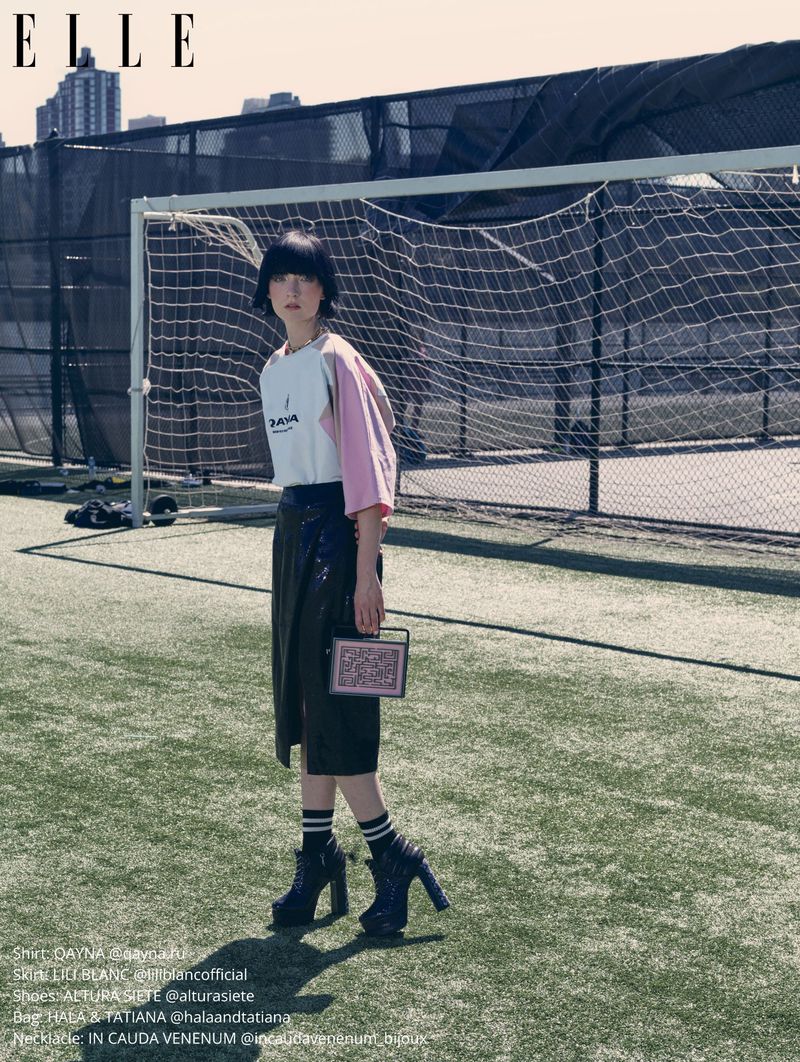 A woman in high-heeled boots stands on a green sports field next to a soccer goal, carrying a small patterned purse, on a sunny day.