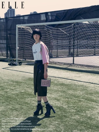 A woman in high-heeled boots stands on a green sports field next to a soccer goal, carrying a small patterned purse, on a sunny day.