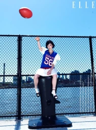 Woman in blue sports jersey and white skirt sitting on a black bollard tossing a red ball against chain-link fence and water view