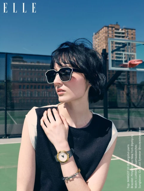A dark-haired woman wearing oversized sunglasses and a black and white top on an outdoor tennis court.