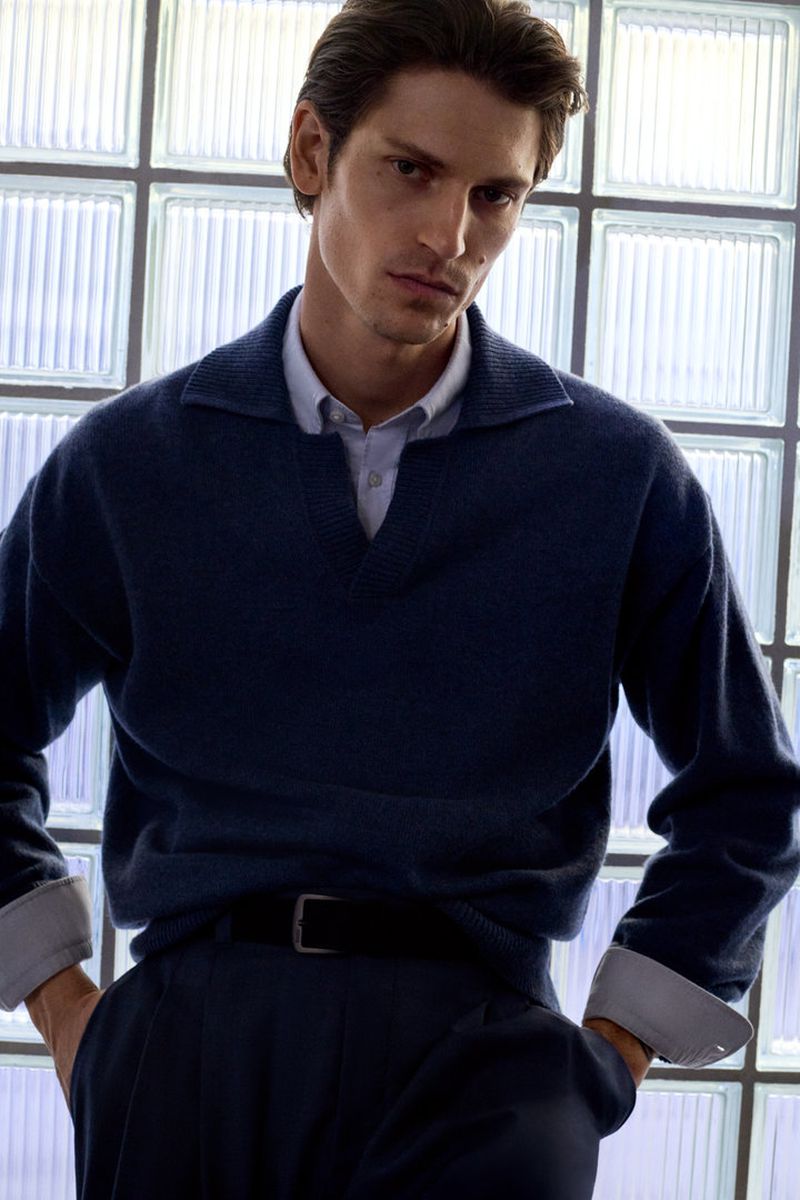 Studio shot of an adult male model wearing a navy-blue sweater and trousers, with a light blue shirt, against a translucent block window backdrop.