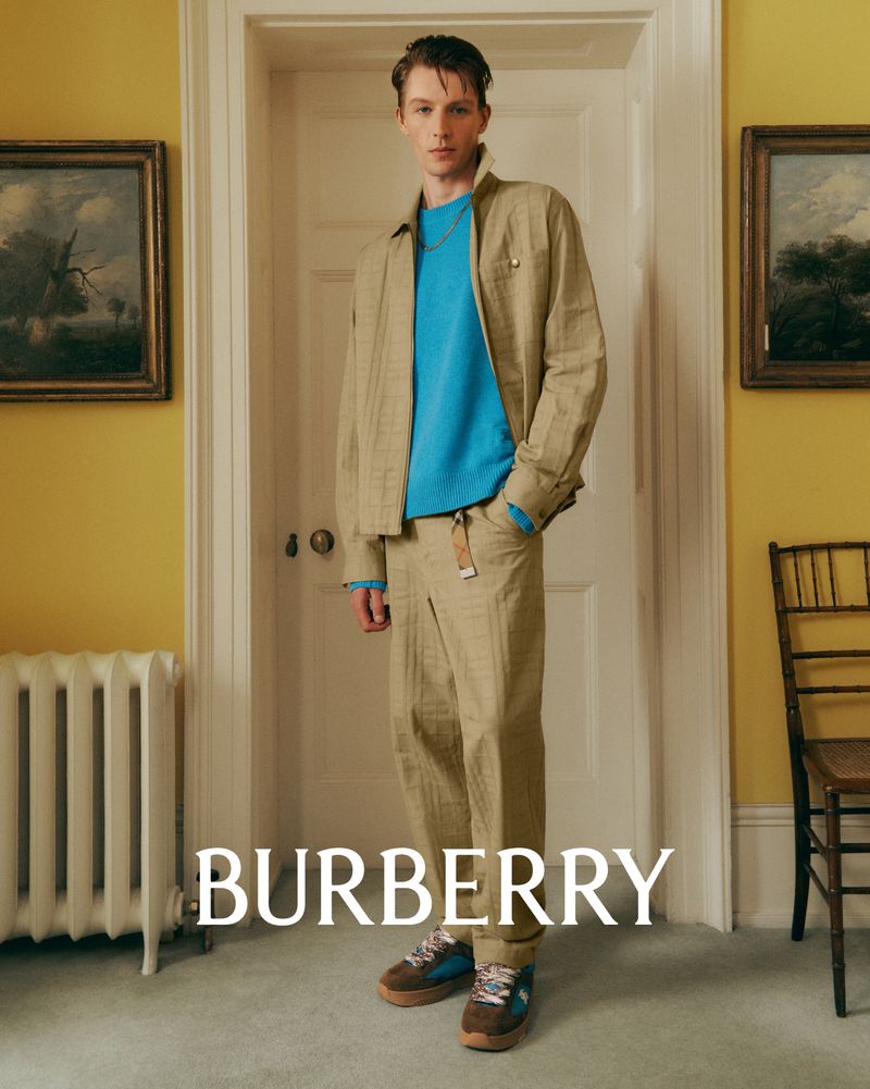 A young adult male poses indoors wearing a khaki jacket and pants with a bright blue sweater, styled with low-top sneakers and gold chain.