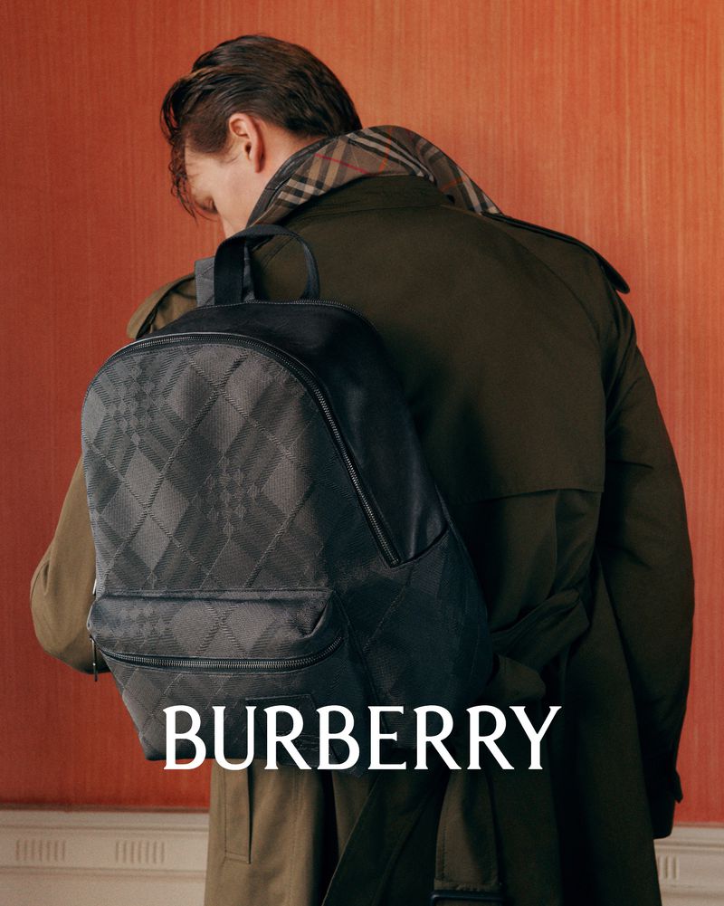 Fashion shot featuring a man with his back to the camera wearing a trench coat and carrying a Burberry backpack indoors.