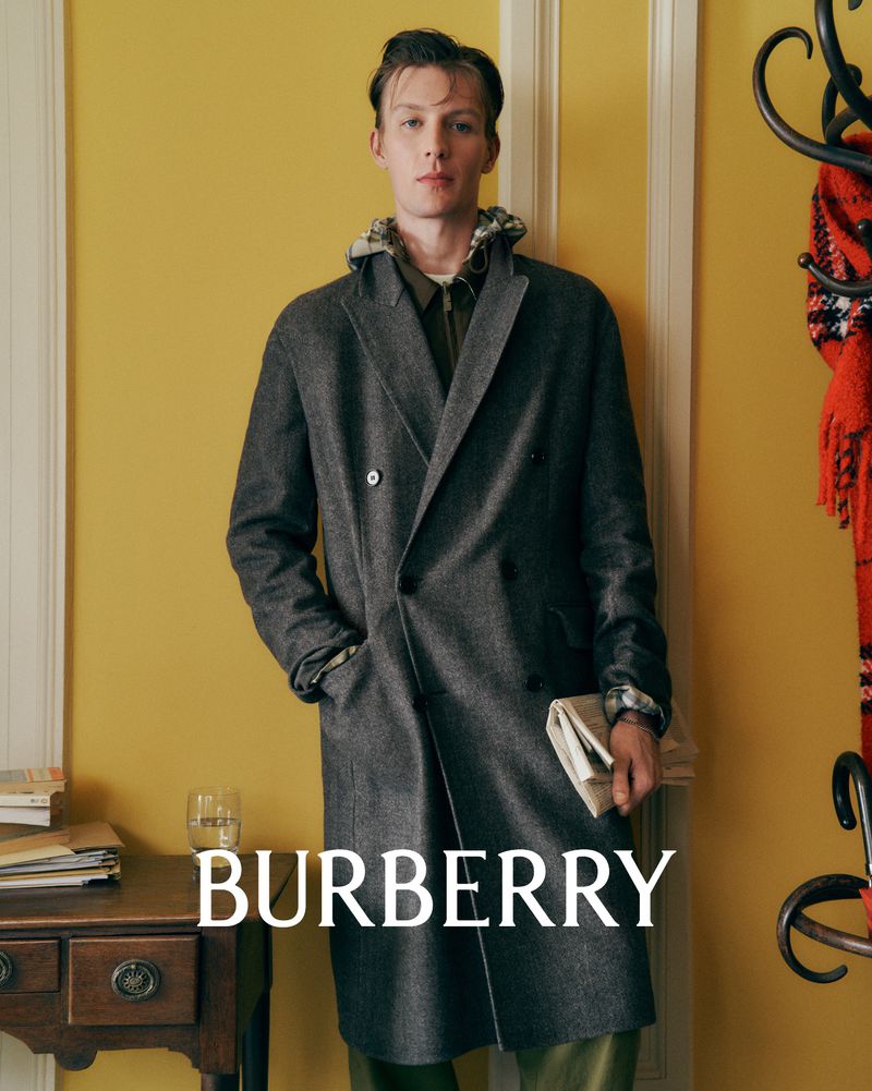 A stylish adult man wearing a grey overcoat poses indoors against a yellow wall, holding a newspaper, in a fashion editorial-style shot.