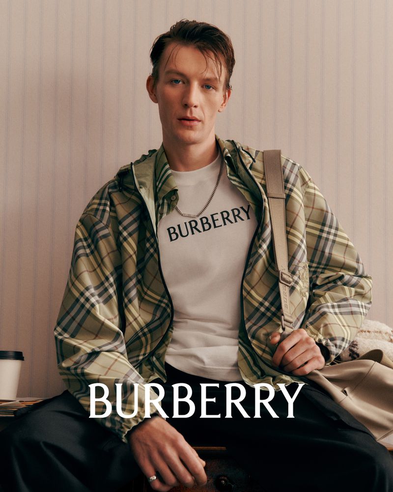 Male model wearing a Burberry t-shirt and jacket sitting indoors with a coffee cup and magazines in a neutral light setting.