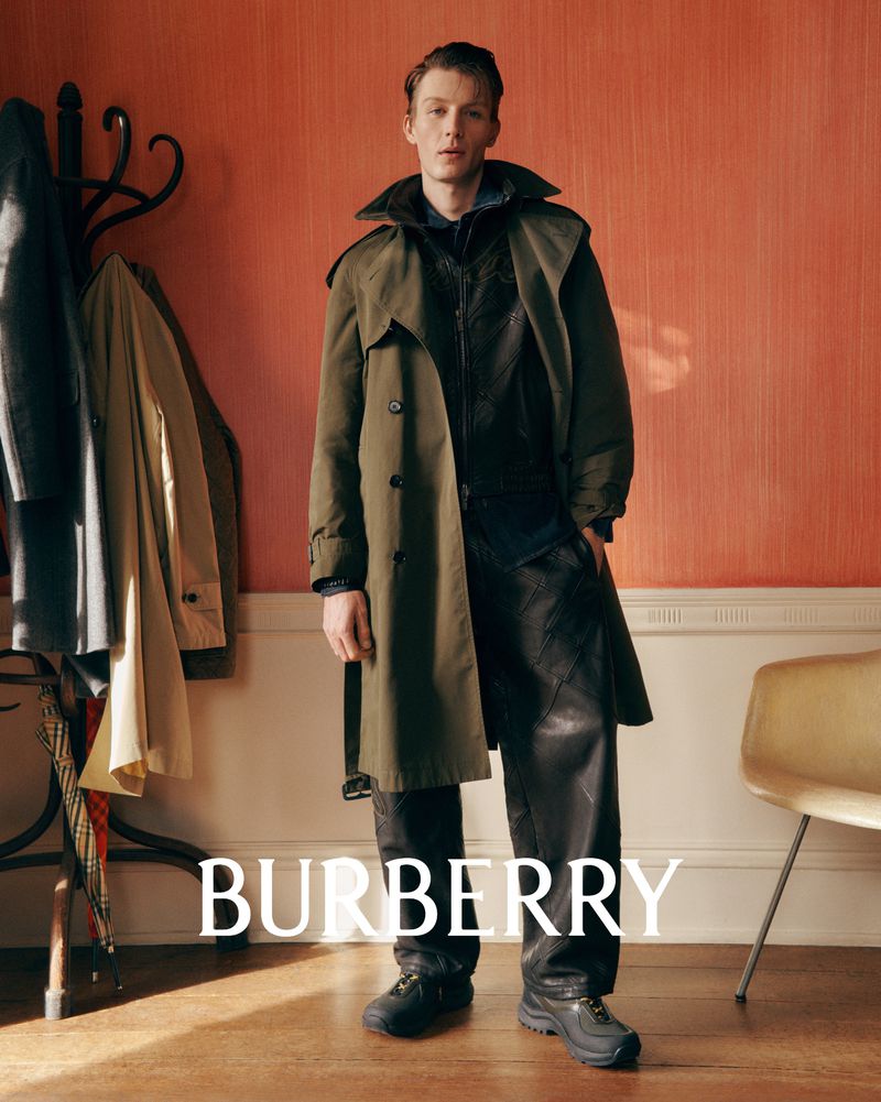 Full-length shot of a man wearing a green trench coat and black leather pants, standing indoors with light walls and Burberry logo.
